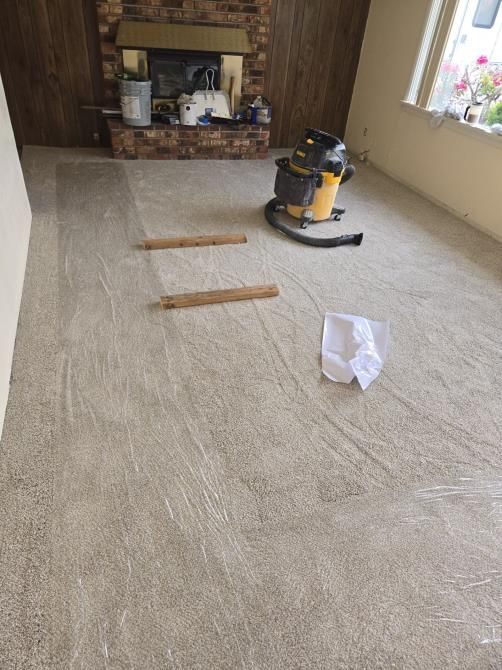 Beige carpet partially installed in a living room; vacuum cleaner and debris present.
