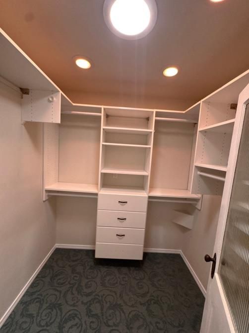 Walk-in closet with white shelves and drawers, beige ceiling, and green carpet.