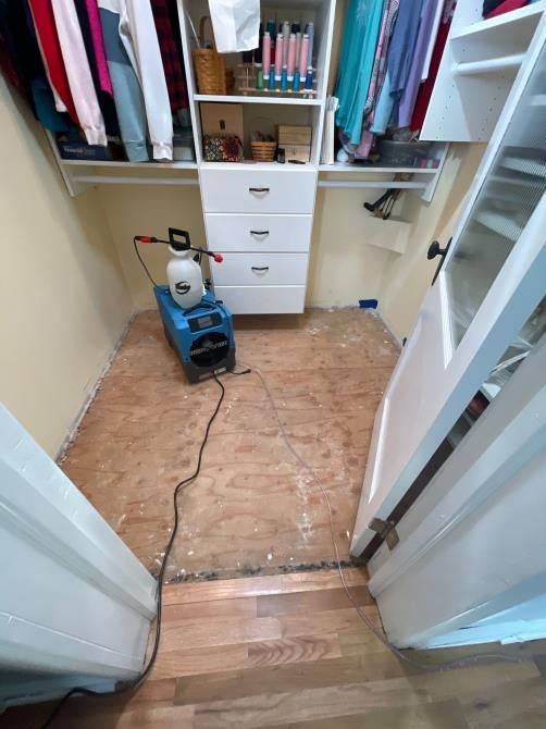 Dehumidifier in a closet with damaged flooring, adjacent to a door, shelves with clothes and drawers.