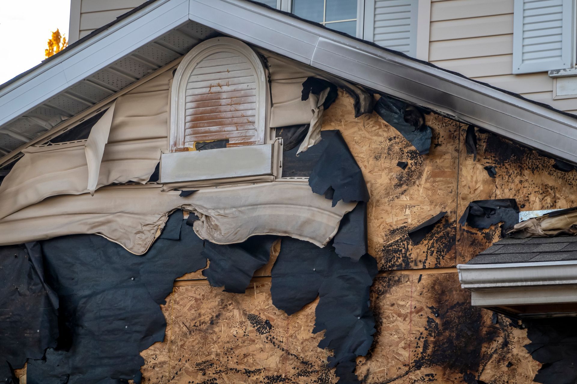 Damaged house siding exposing plywood and a small window.