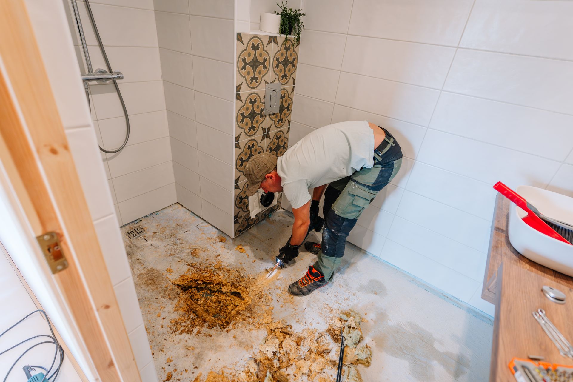 A worker in a bathroom is removing debris from the floor. White walls, patterned tile, and shower fixture.