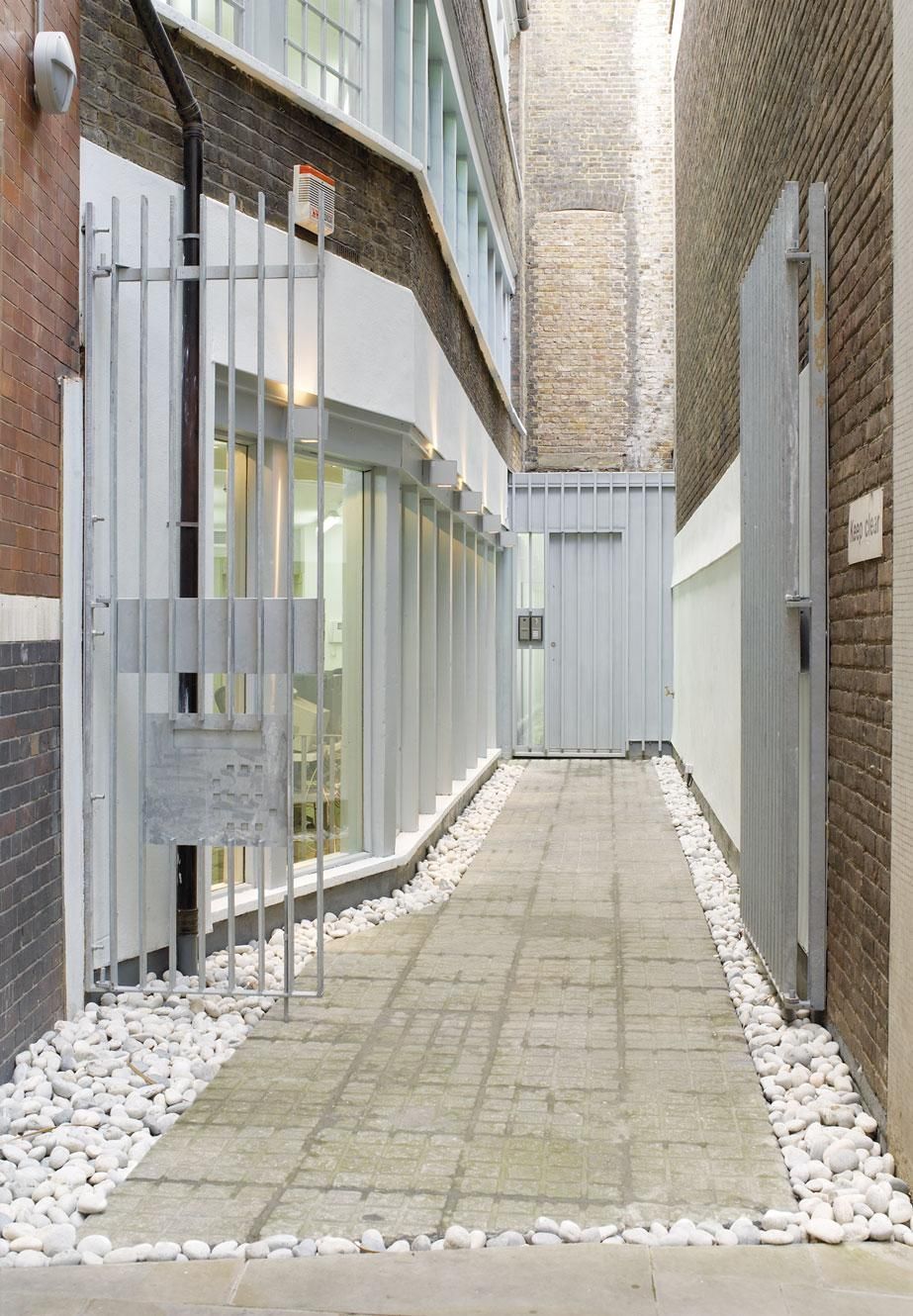 Narrow paved alley between brick buildings, with glass windows, white fence, and white stones along the edges.