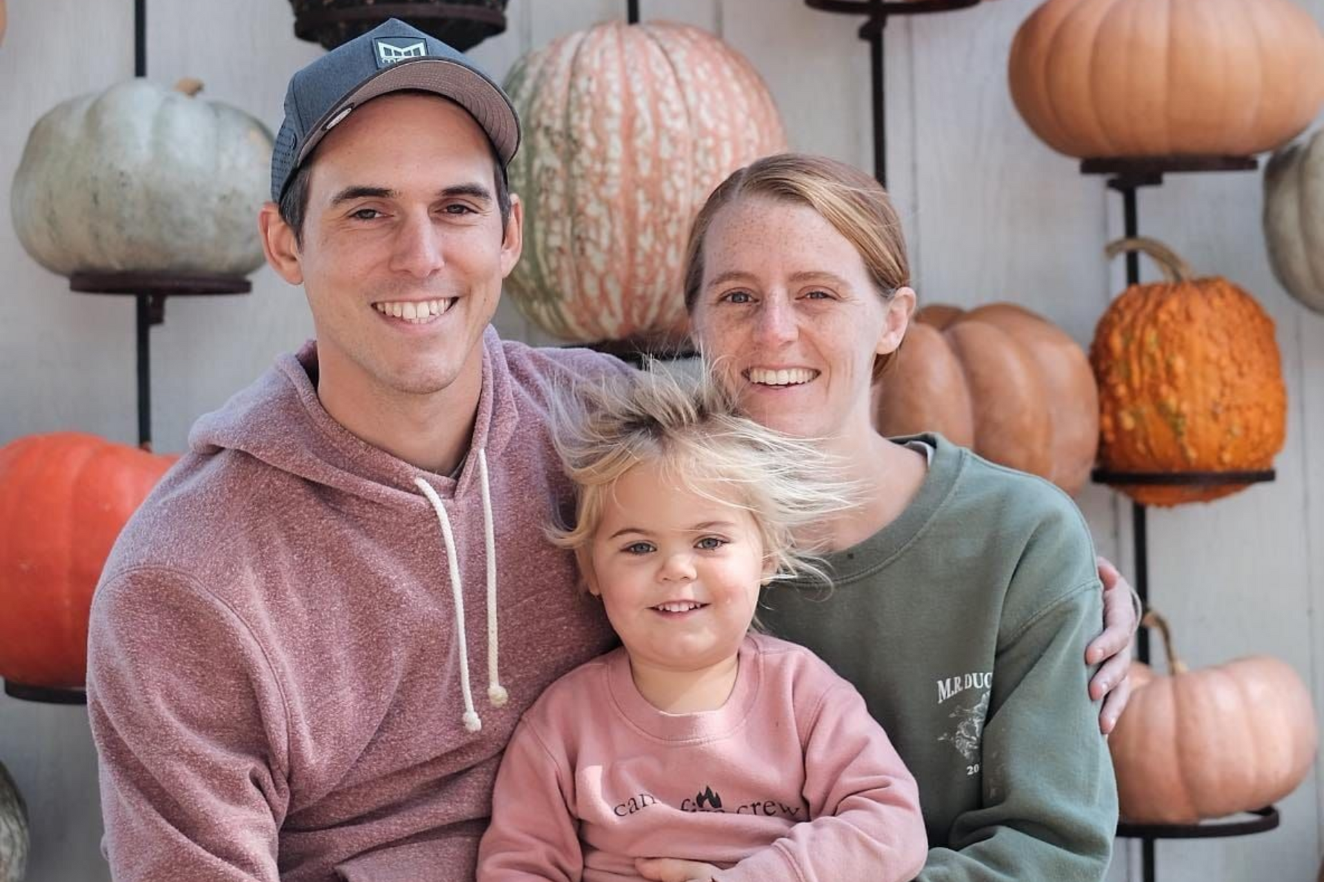 Family smiling in front of pumpkins: father in cap, mother, and child with windswept hair.