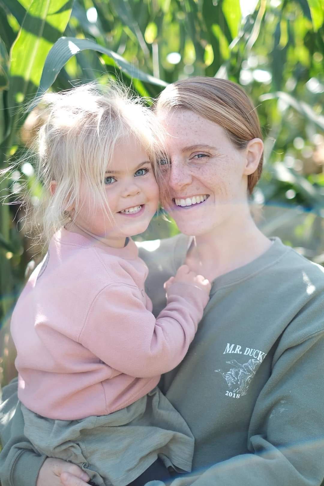 Woman holding a smiling toddler in a cornfield; both looking at camera, sunny day.