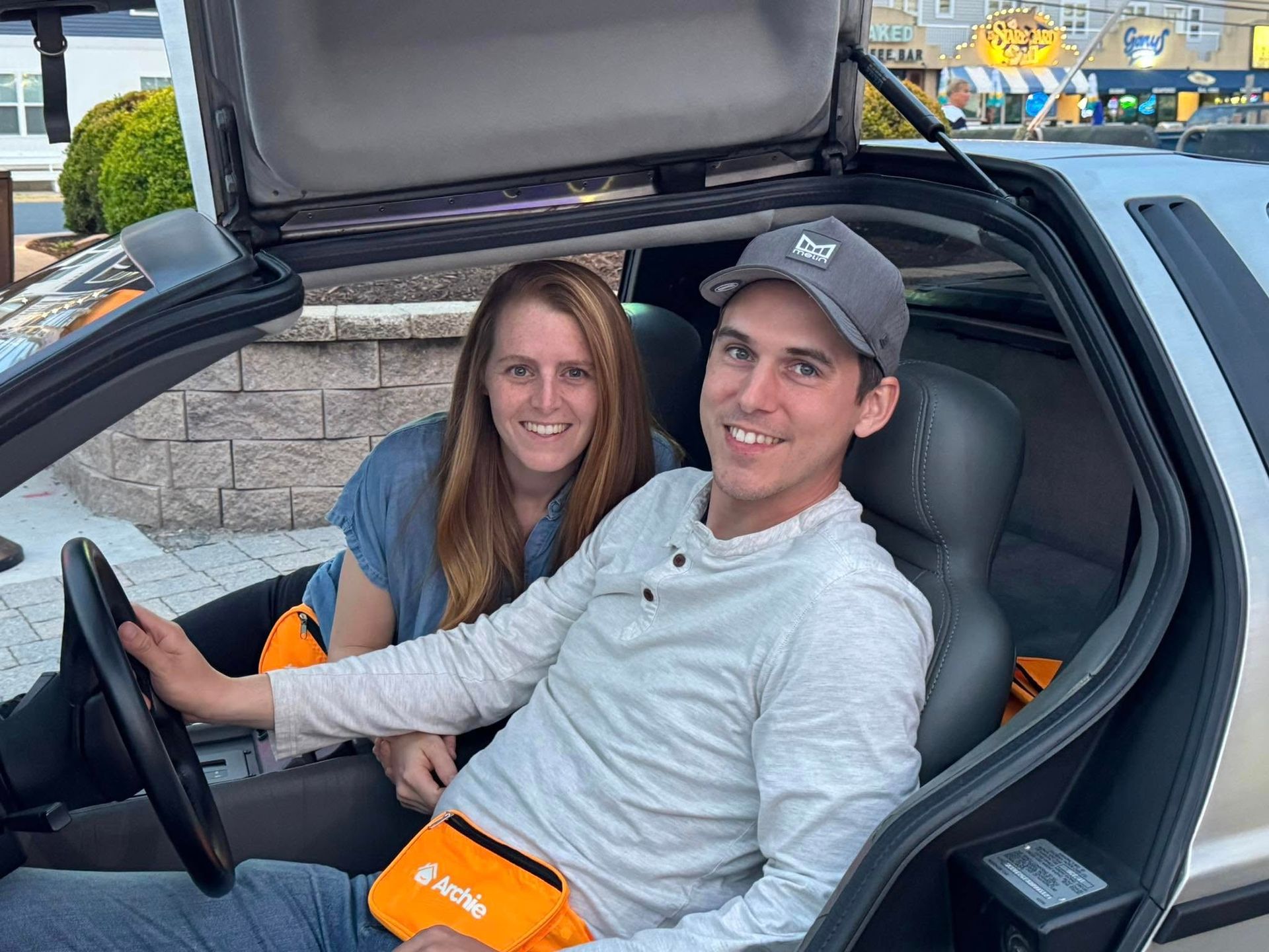 Couple in a DeLorean, smiling, driver holding steering wheel, door open, gray car interior, outdoor setting.
