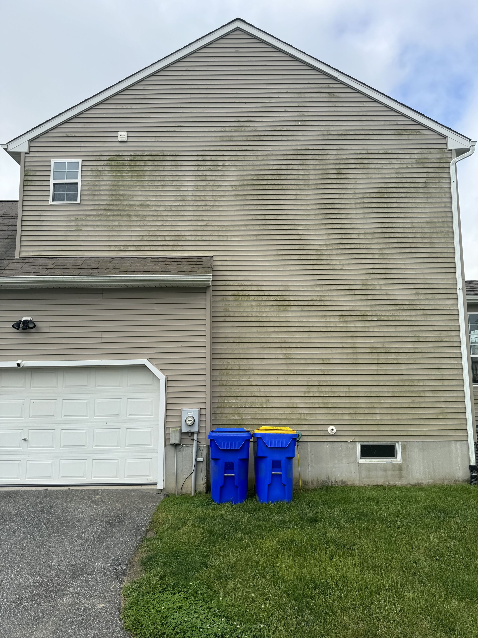 Gray siding of a house with visible green algae, blue trash cans, and a white garage door.