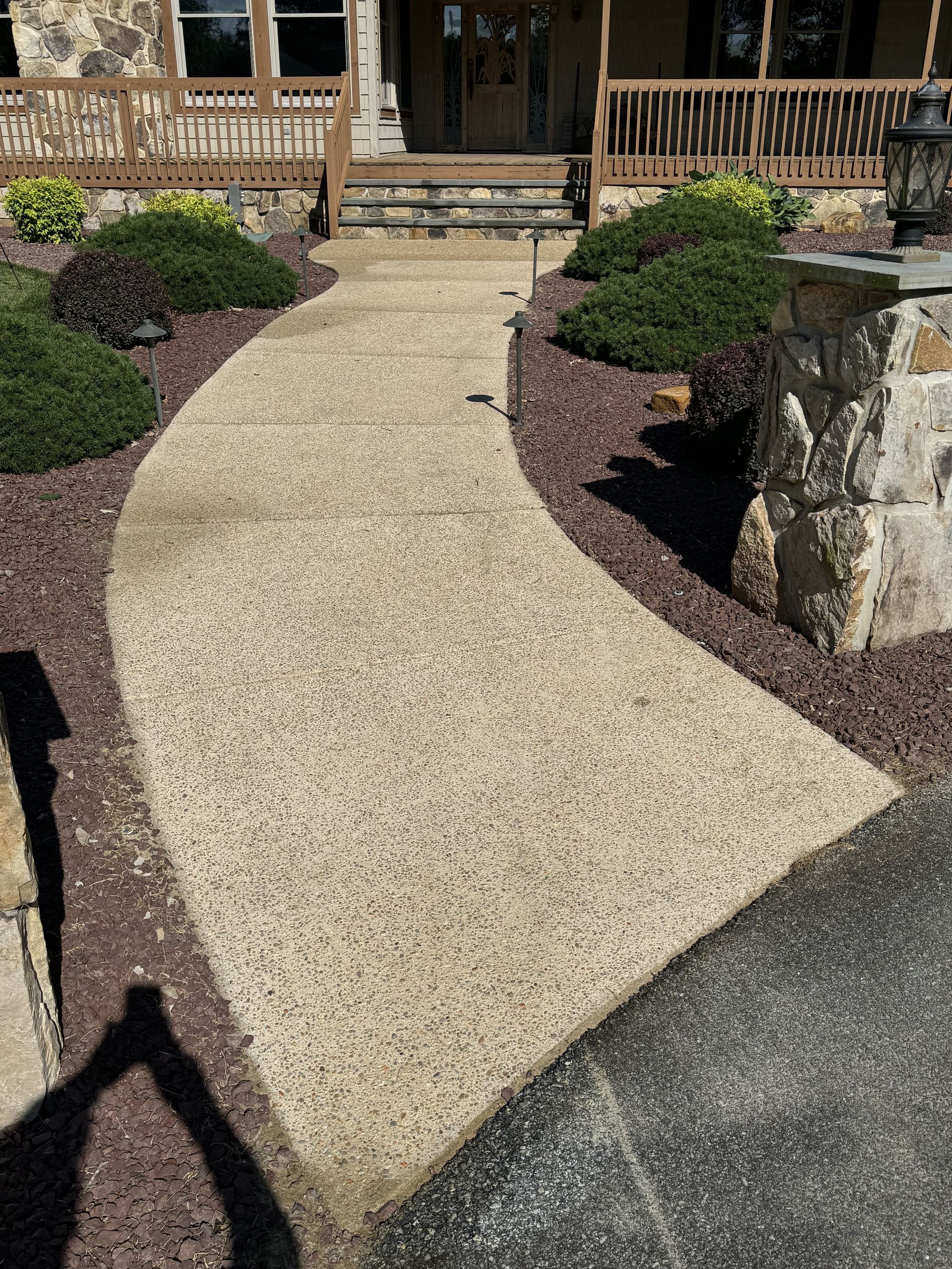 Curving concrete walkway leading to a house entrance, edged with brown mulch and green bushes.