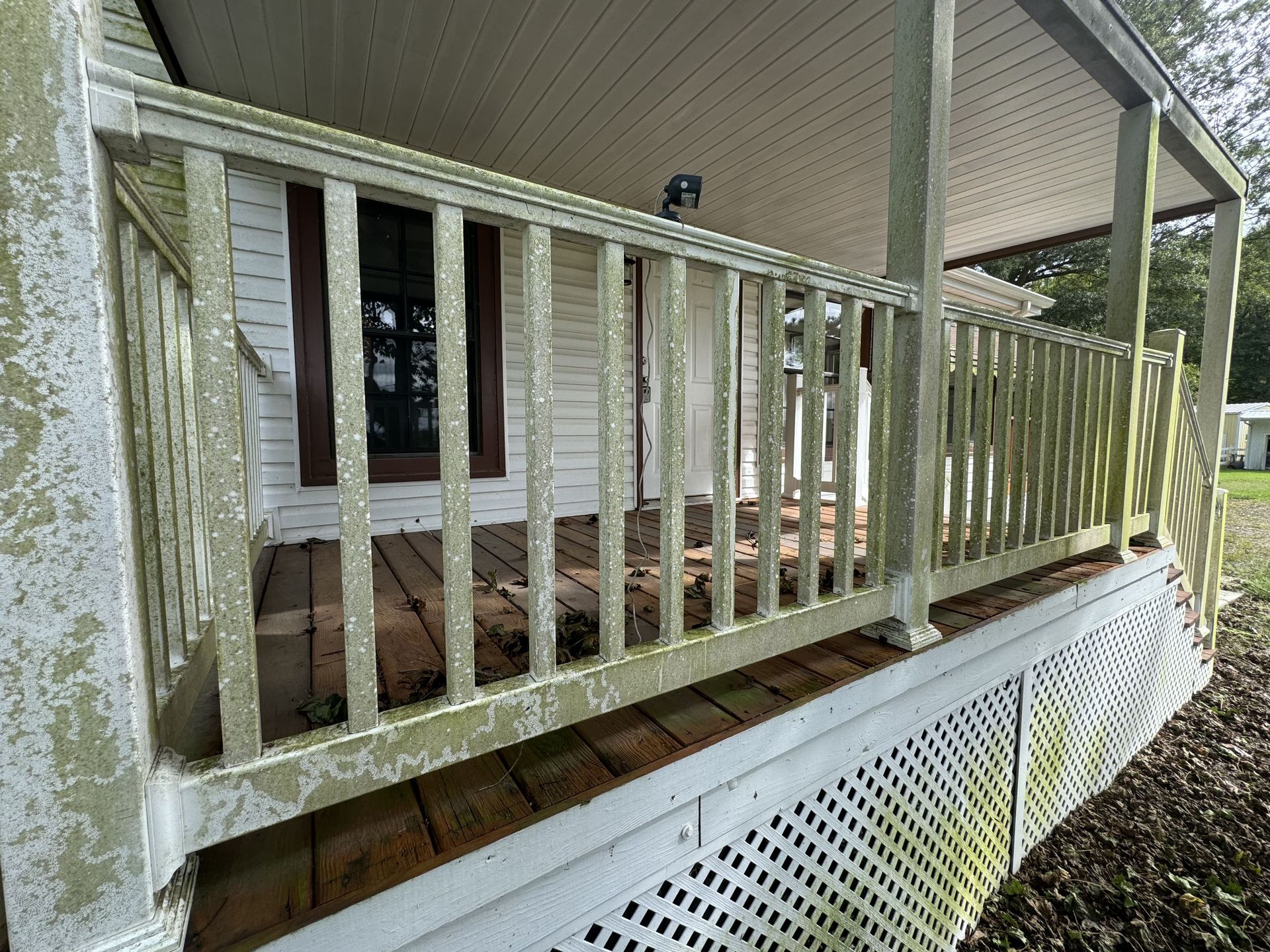 A porch with weathered, green-painted railings, wooden planks, and latticework; white house in the background.