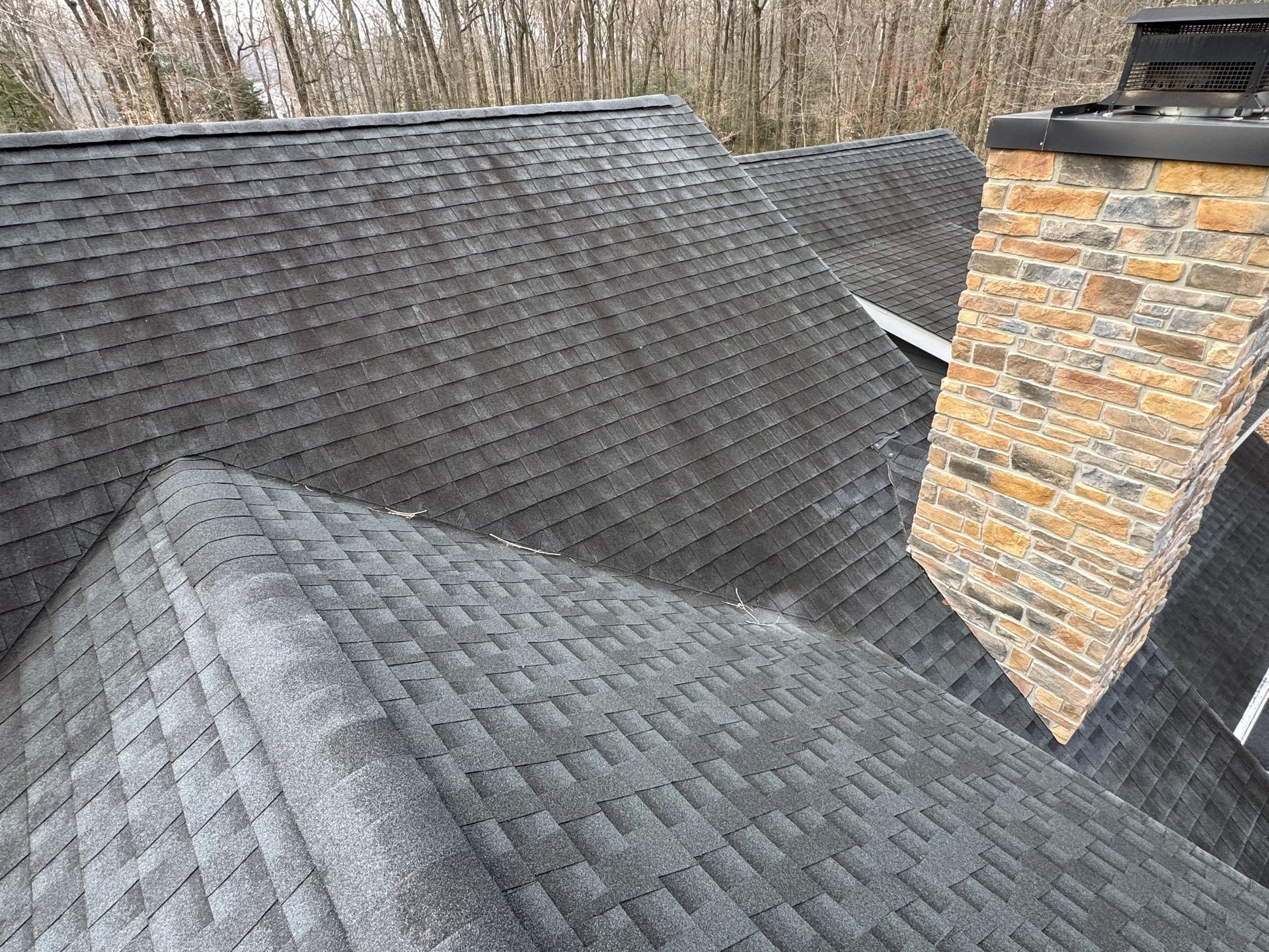 Gray asphalt shingle roof with dark streaks, next to a stone chimney, trees in background.