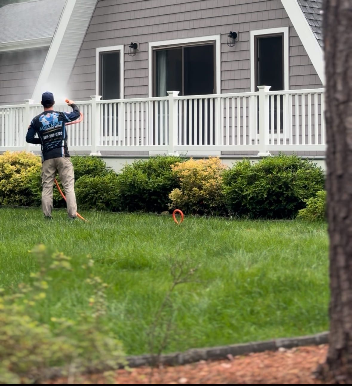 Man power washing the exterior of a gray A-frame house with a white porch. Green bushes and grass surround.