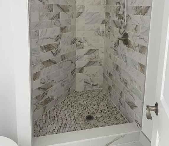 Shower stall with gray and white marble-like tiles on walls and small stone tile floor.