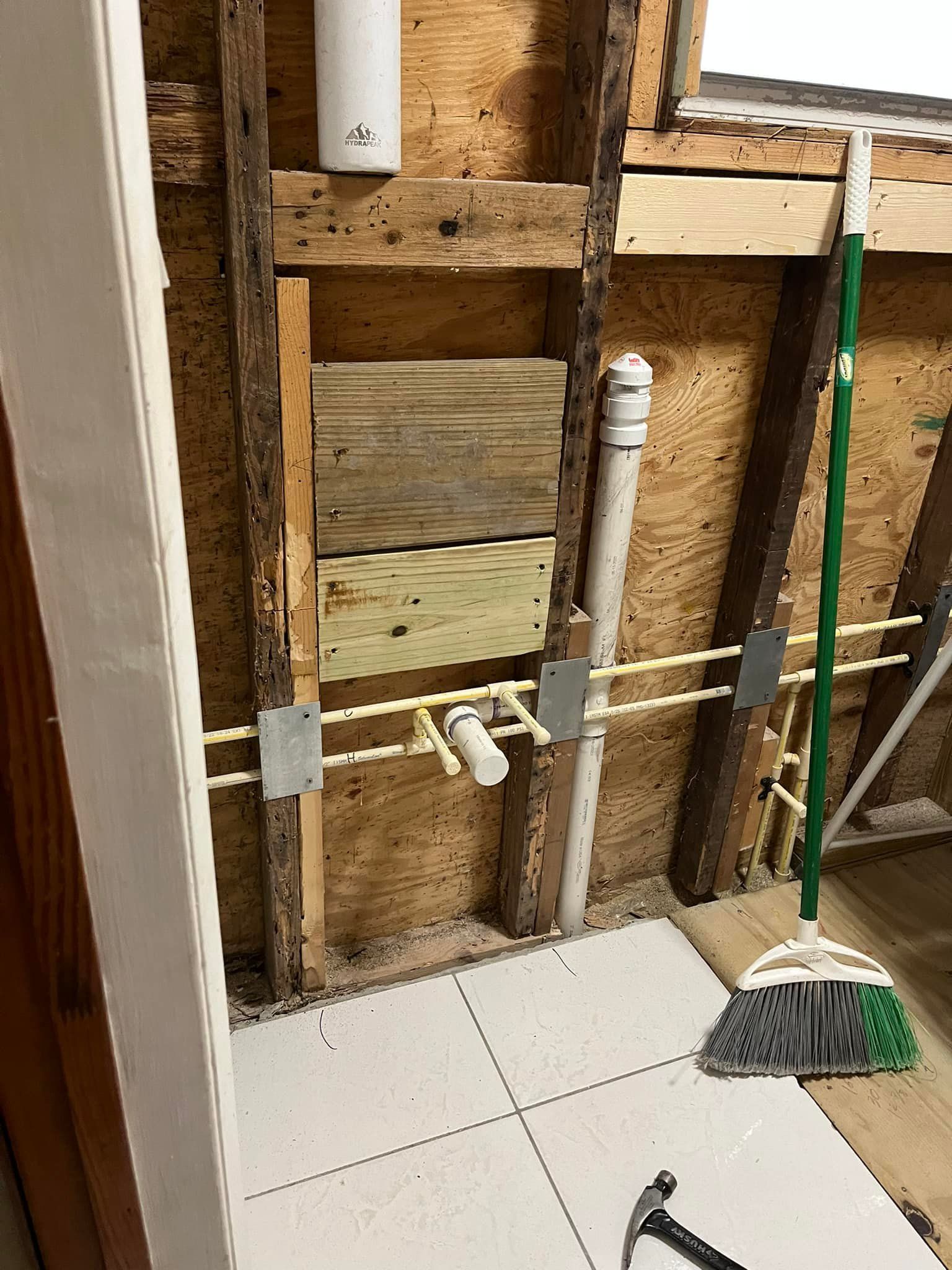 Bathroom wall under construction with exposed plumbing and broom.