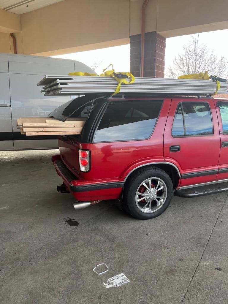 Red SUV with lumber on roof rack and in back, parked under a shelter.