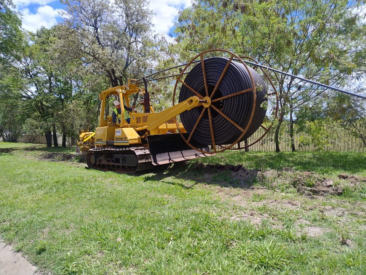 Um trator de esteiras amarelo com um grande carretel de cabo preto montado na traseira, em um campo gramado e arborizado.