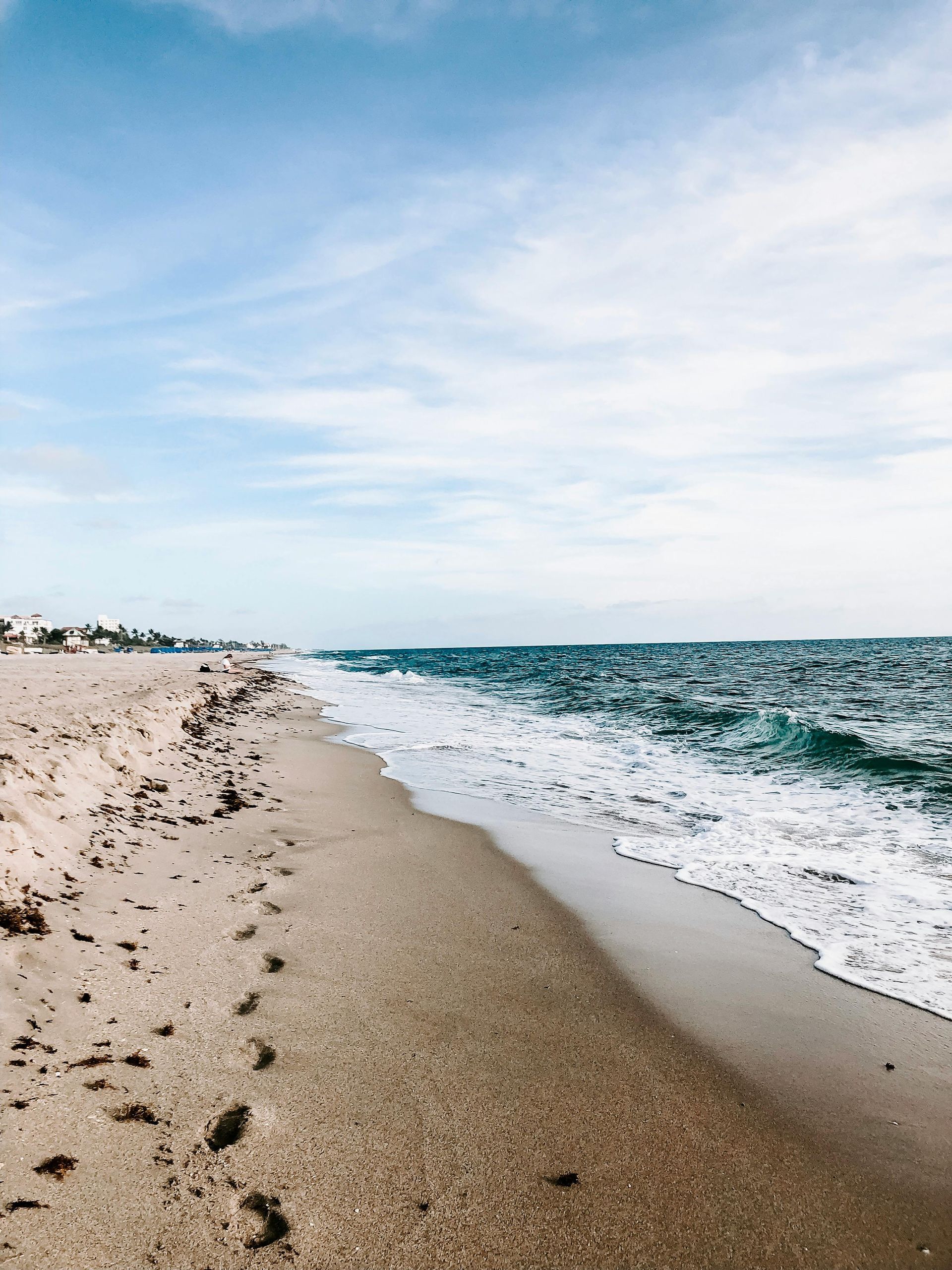 Sandy beach meets ocean under a blue, cloudy sky. Footprints in the sand.