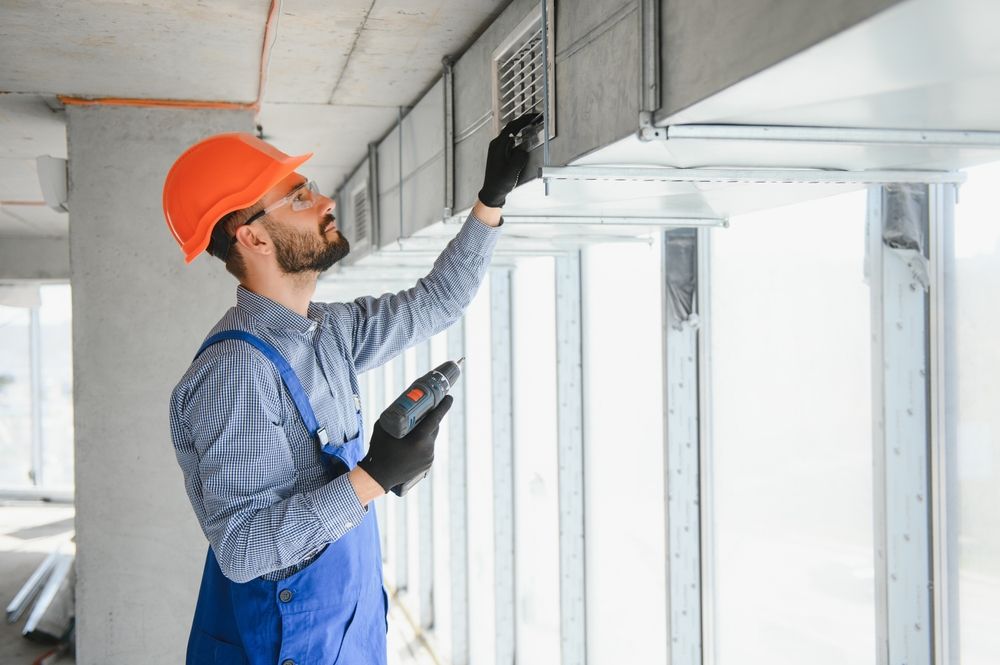 Construction worker in orange hard hat inspecting ductwork with a tool.