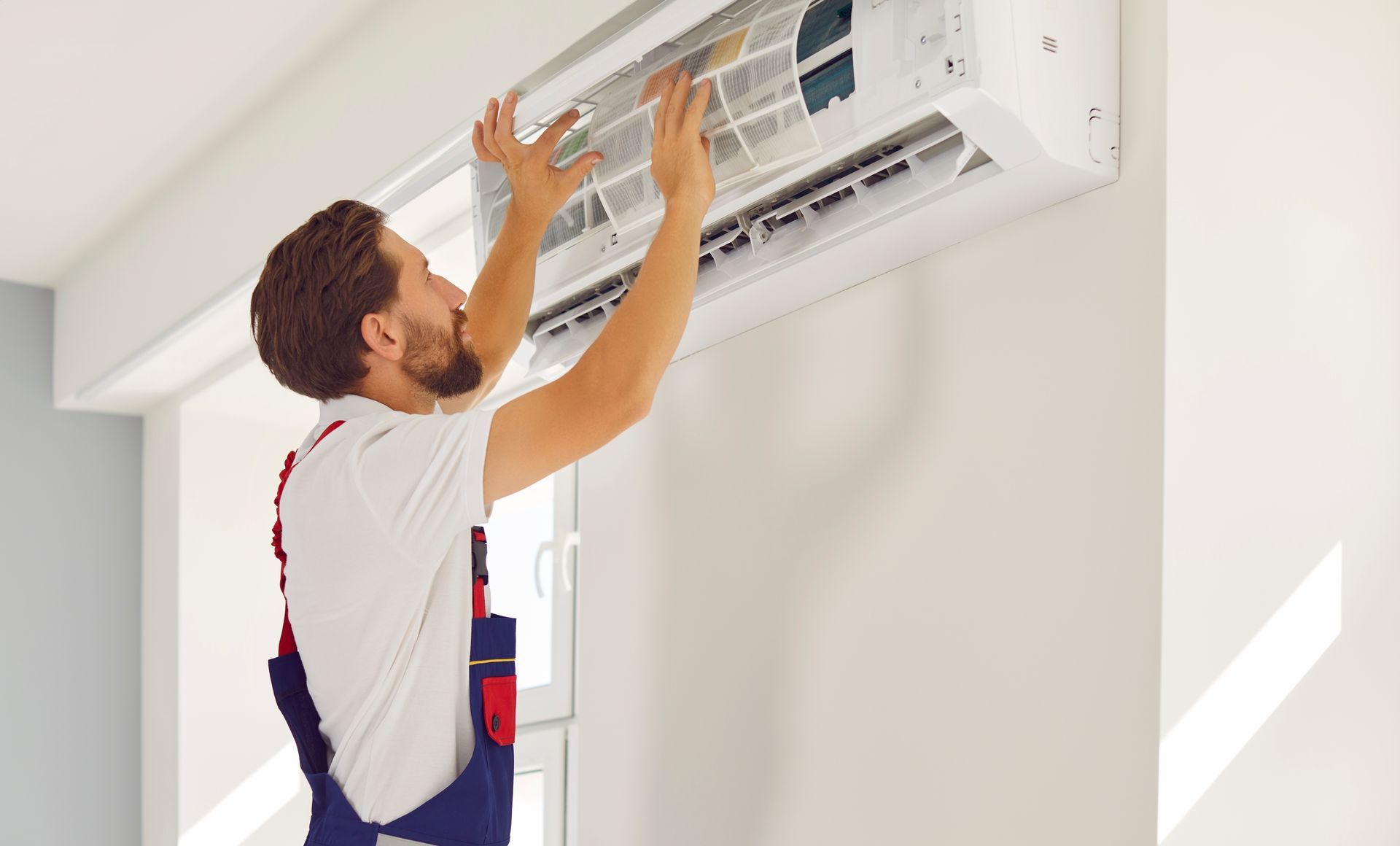 A person in overalls cleans the filter of a white air conditioning unit mounted on a wall.