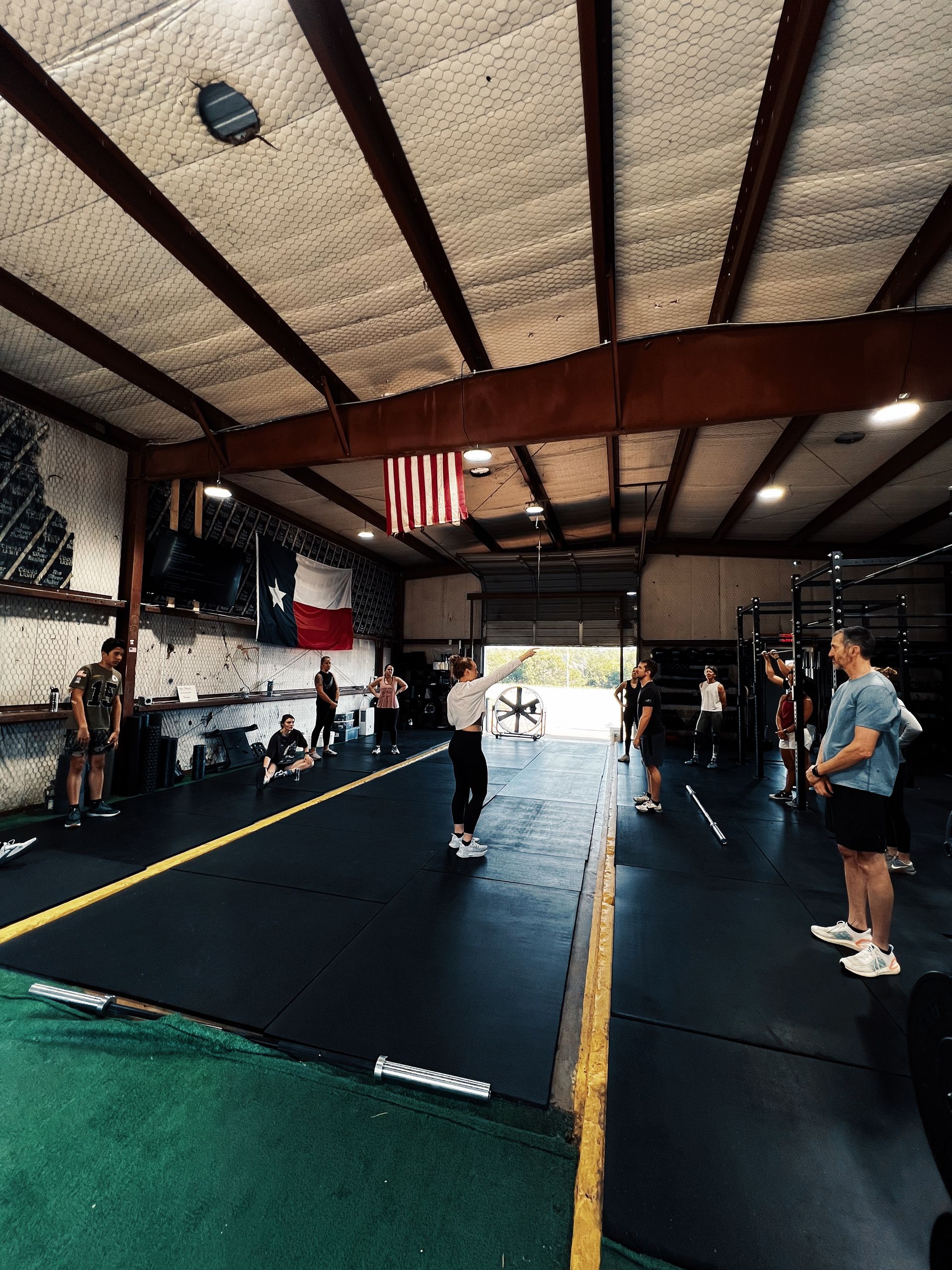 A group of people are standing in a gym with an american flag hanging from the ceiling.