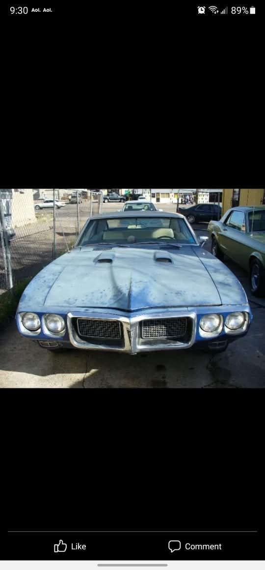A blue vintage Pontiac Firebird parked in a lot, viewed from the front, showing weathered paint and dual hood scoops.