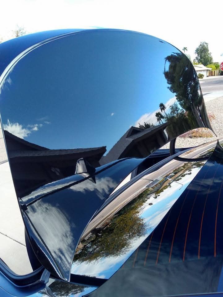 A reflective, black car trunk lid angled open, showing the mirror-like reflection of a house, sky, and trees.