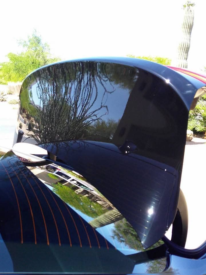The glossy, dark-painted trunk lid of a car is open, reflecting the surrounding trees and buildings outdoors.