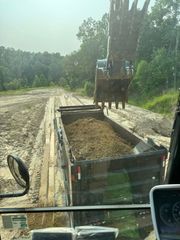 An excavator loading a dump truck with dirt in a muddy field with trees in the background.