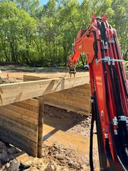 Orange excavator working on a construction site with wooden beams and a muddy ground. Trees in the background.
