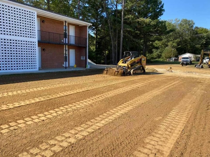 A skid steer levels a dirt lot next to a brick building on a sunny day.