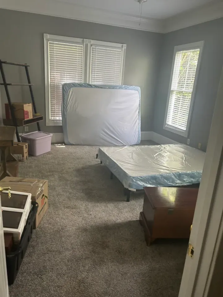 A bedroom with gray walls, carpeted floor, a bed frame, a mattress against a window, storage boxes, and furniture.