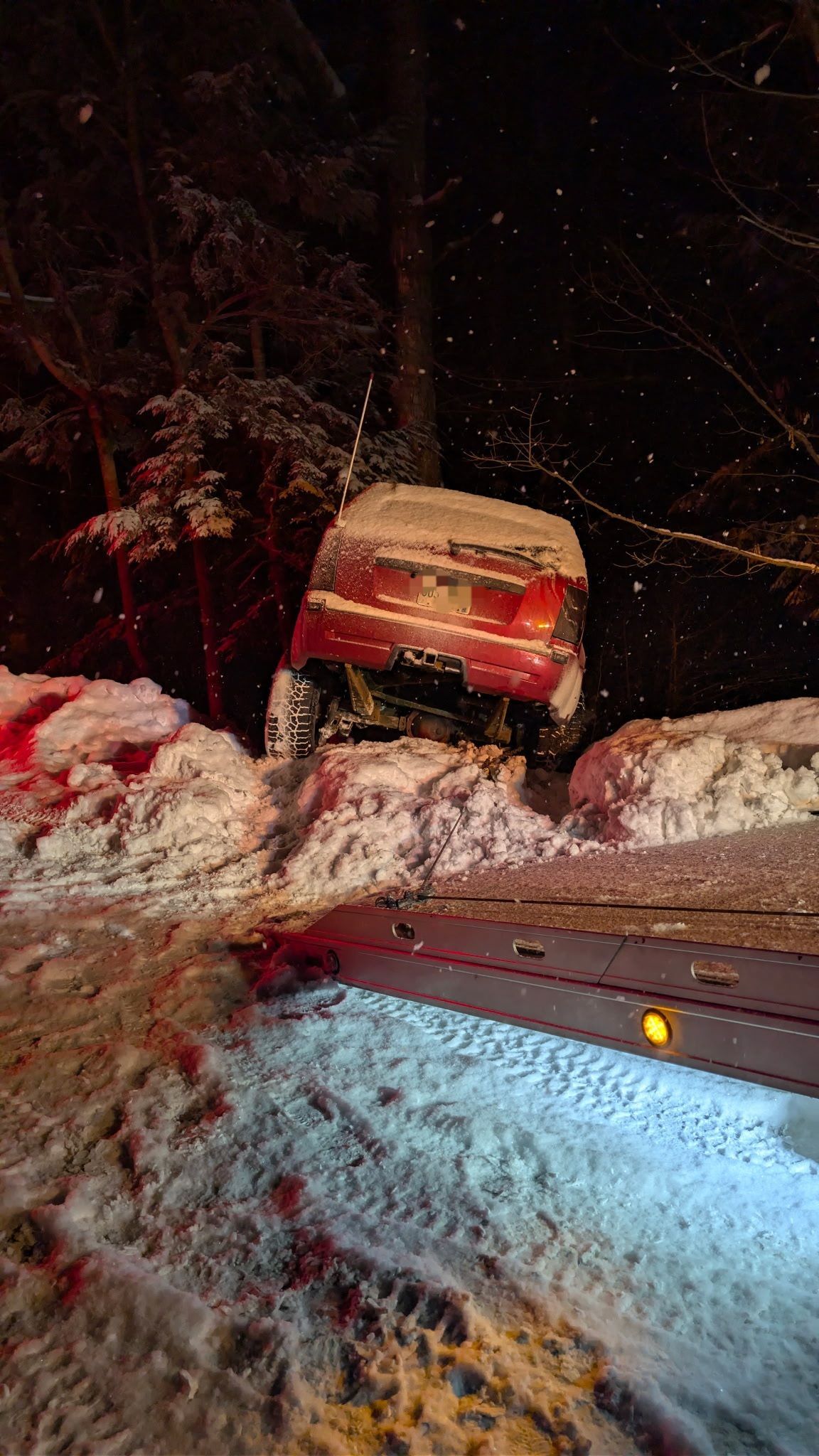 A red vehicle stuck in a snowbank at night, viewed from the rear, with a tow truck bed visible in the foreground. MAACS Tow LLC
