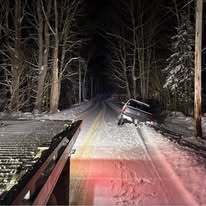 A car is stuck off the side of a snowy, dark road at night, viewed from the perspective of a tow truck. MAACS Tow LLC