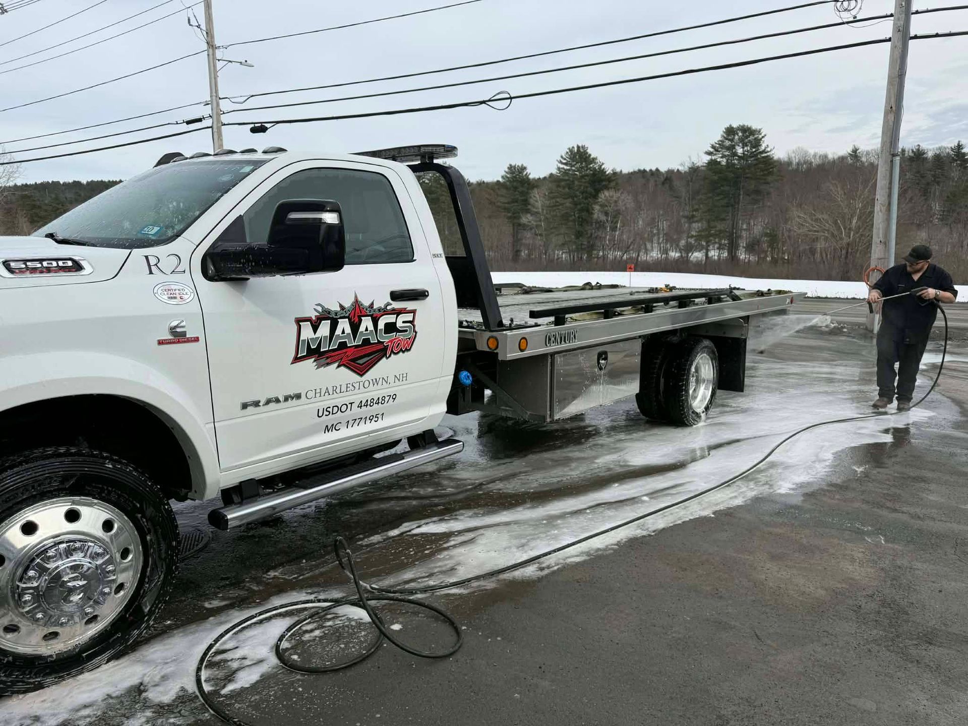 A person uses a pressure washer to clean a white flatbed tow truck parked on an asphalt lot in a wooded setting. MAACS Tow LLC