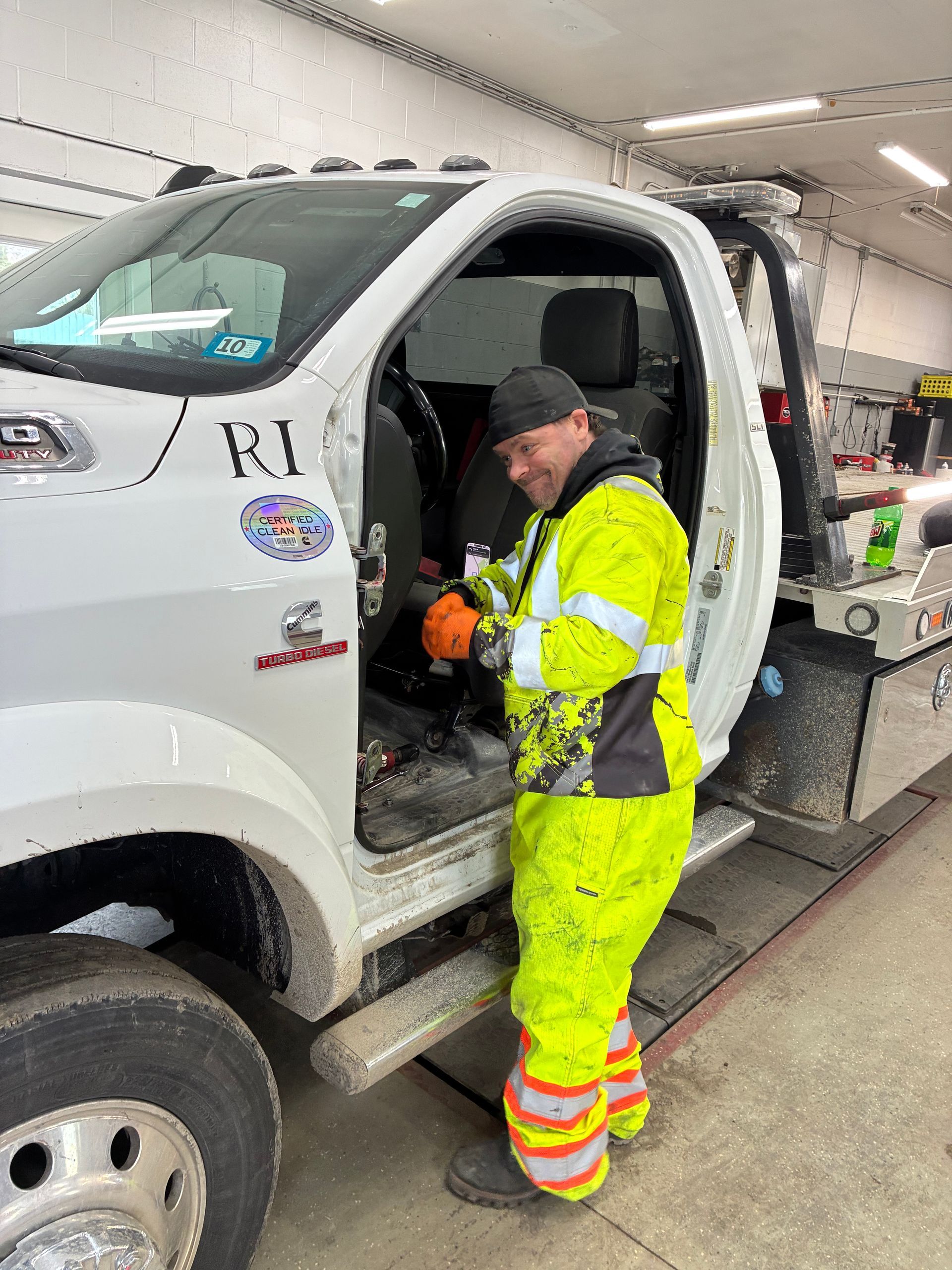 A person in high-visibility neon work gear smiles while stepping out of a white tow truck inside a garage. MAACS Tow LLC