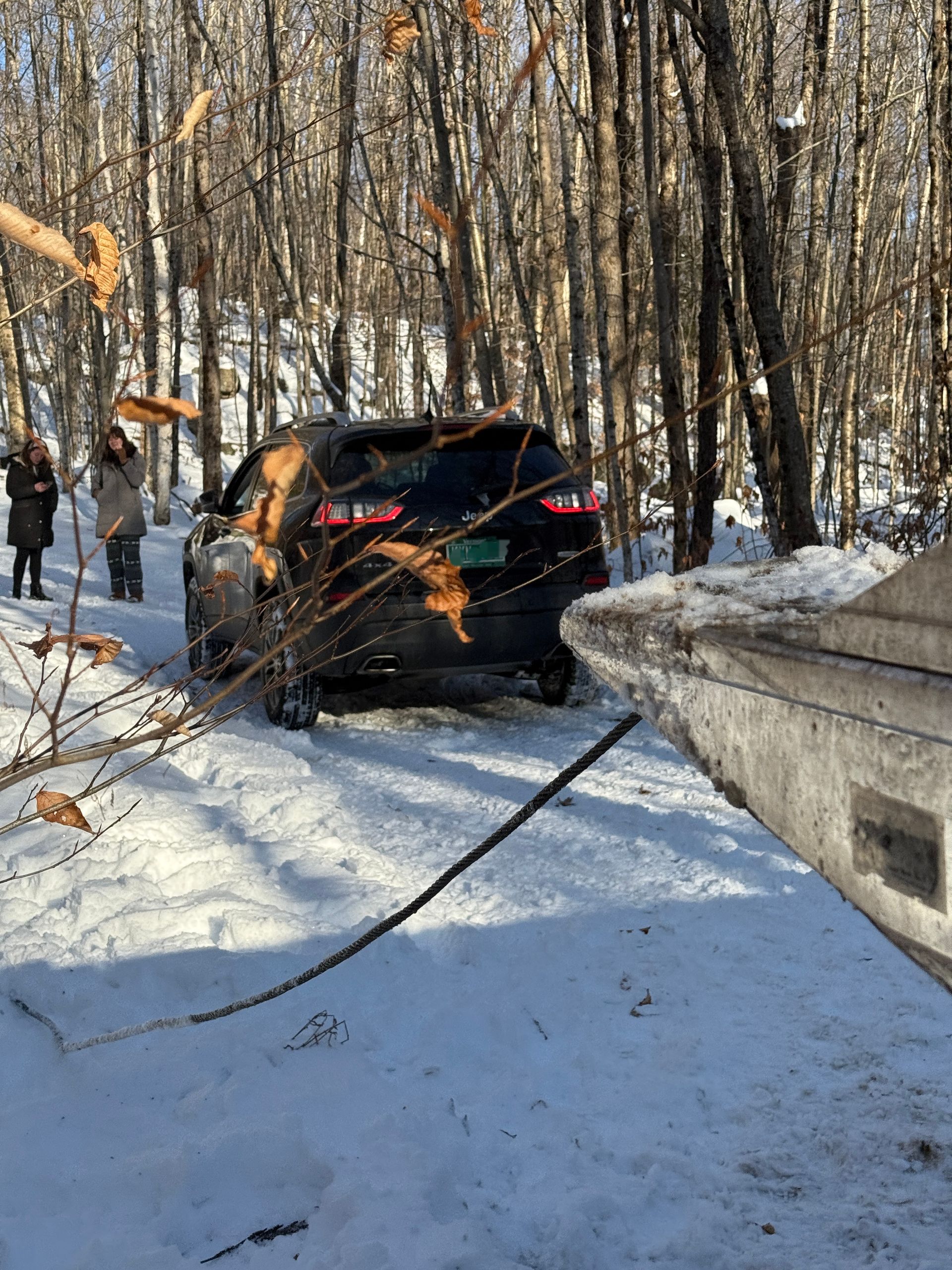 A dark SUV stuck in deep snow in a woods-lined area, with a tow cable connected to it from the foreground. MAACS Tow LLC