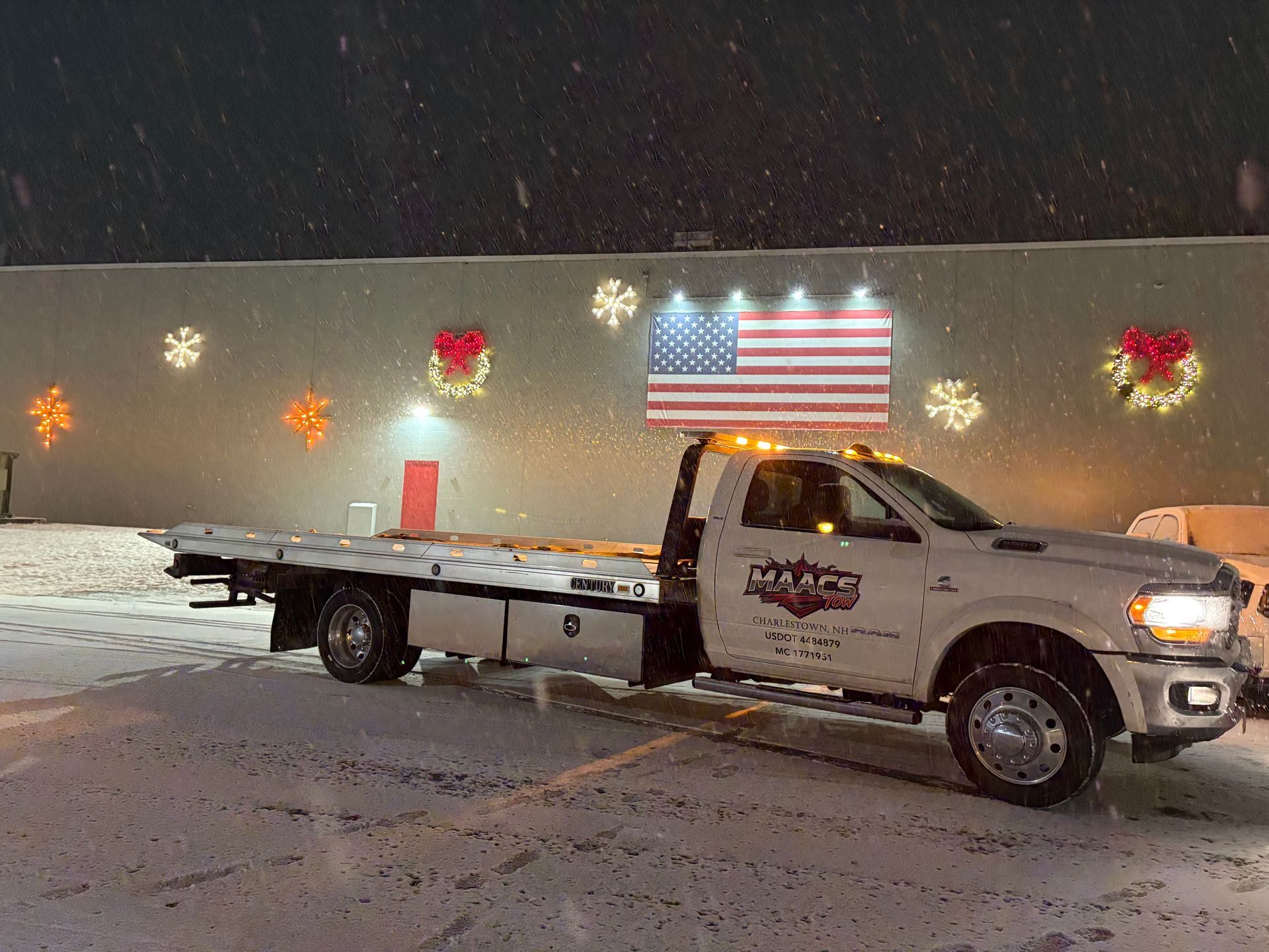 A white flatbed tow truck parked in a snowy lot at night, featuring a prominent illuminated American flag on its roof. MAACS Tow LLC