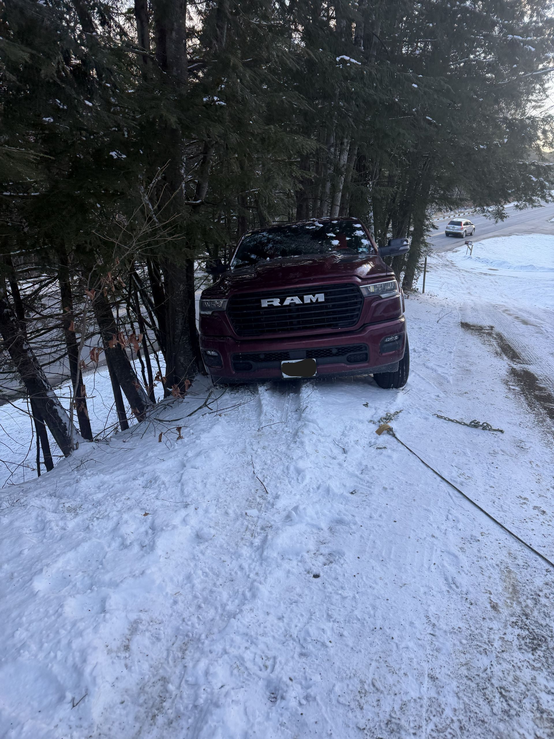 A maroon RAM pickup truck stuck off a snowy road, positioned near a cluster of pine trees. MAACS Tow LLC