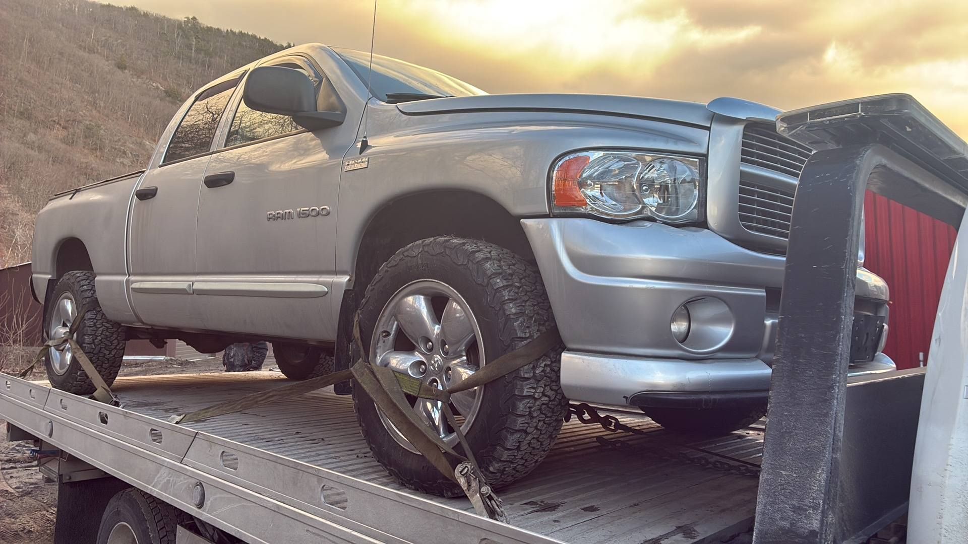 A silver Dodge Ram pickup truck is secured with straps on a flatbed tow truck at sunset. MAACS Tow LLC