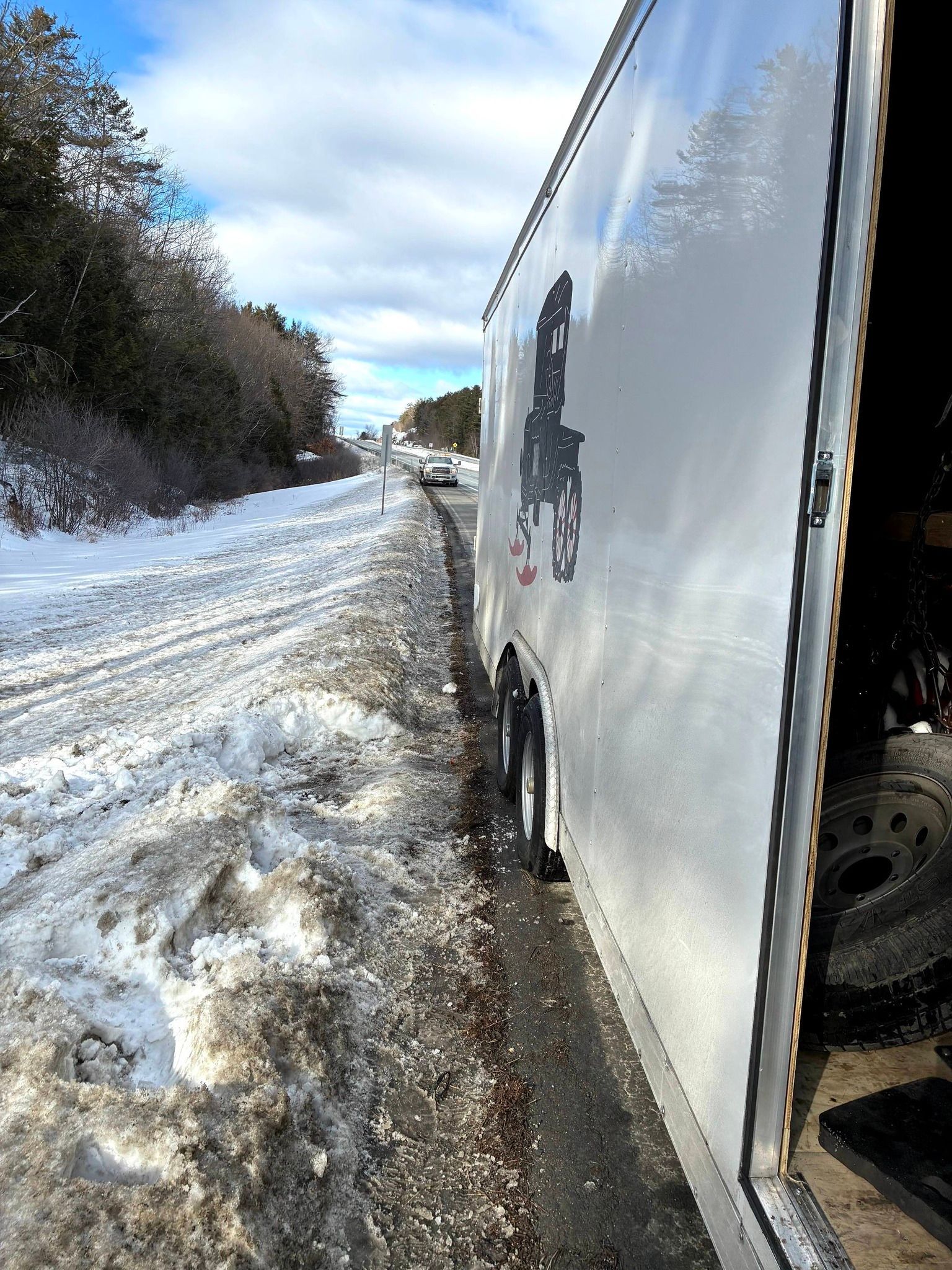 A white cargo trailer parked on the snowy shoulder of a highway, viewed from the side. MAACS Tow LLC