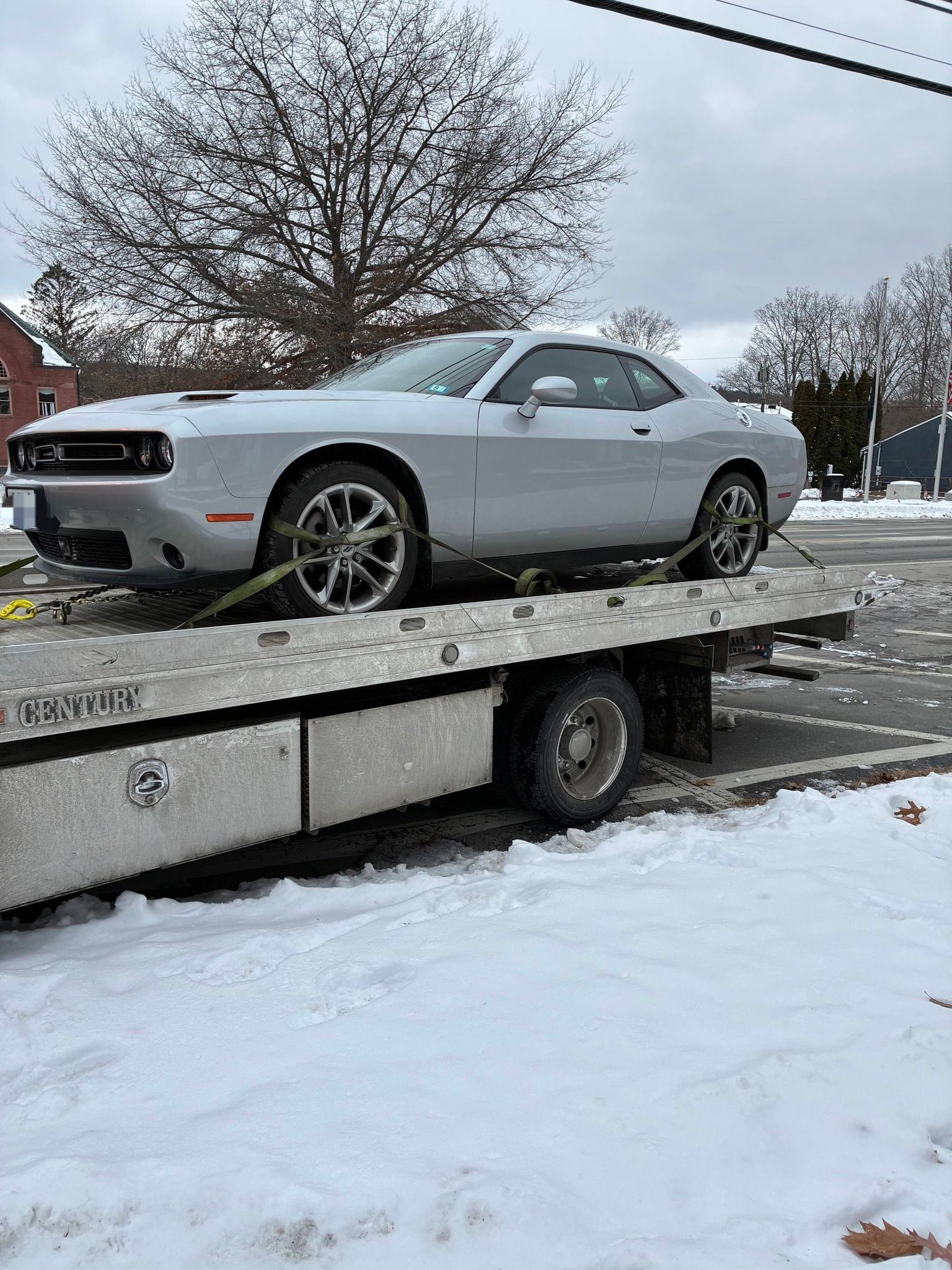 A silver Dodge Challenger loaded onto a flatbed tow truck on a snowy, overcast day. MAACS Tow LLC