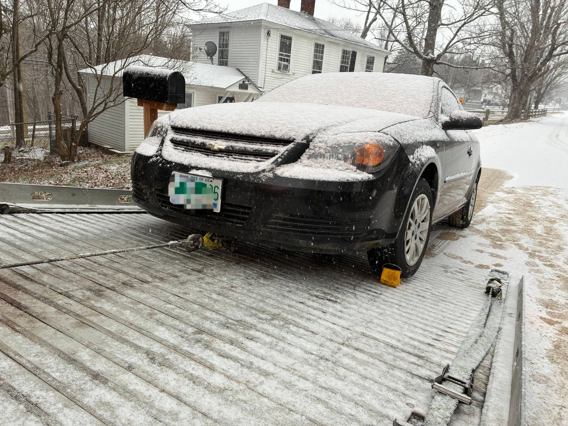 A black sedan covered in a light dusting of snow is secured with yellow wheel chocks on the metal bed of a tow truck. MAACS Tow LLC