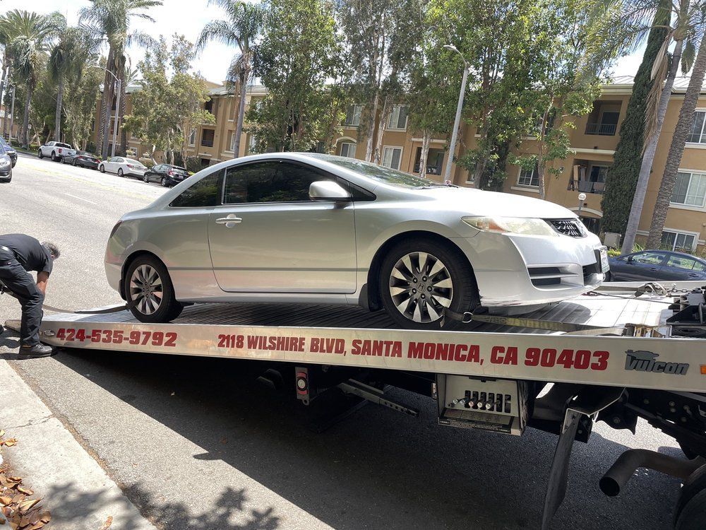 Silver car being towed onto a flatbed tow truck on a street. A person works on the truck.