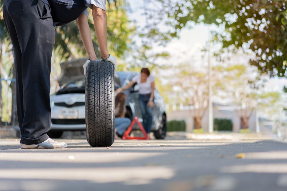 Person holding a tire, car with open hood and a woman near an emergency triangle on a sunny road.