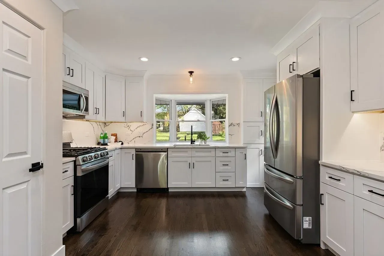 White kitchen with stainless steel appliances, dark wood floors, and a window overlooking a yard.