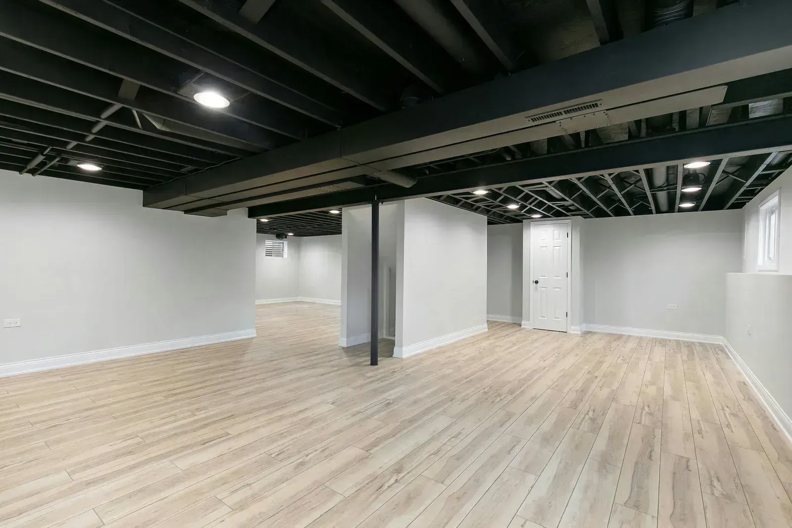 Finished basement with light wood-look flooring, white walls, and black ceiling beams.