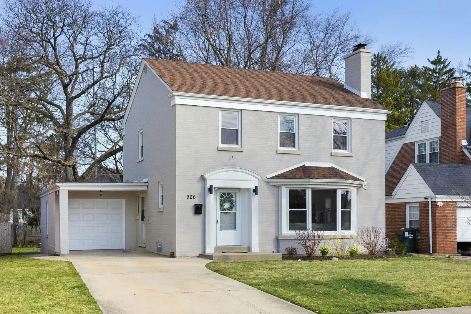 Two-story gray brick house with a brown roof and attached garage, with a curved bay window and a white front door.