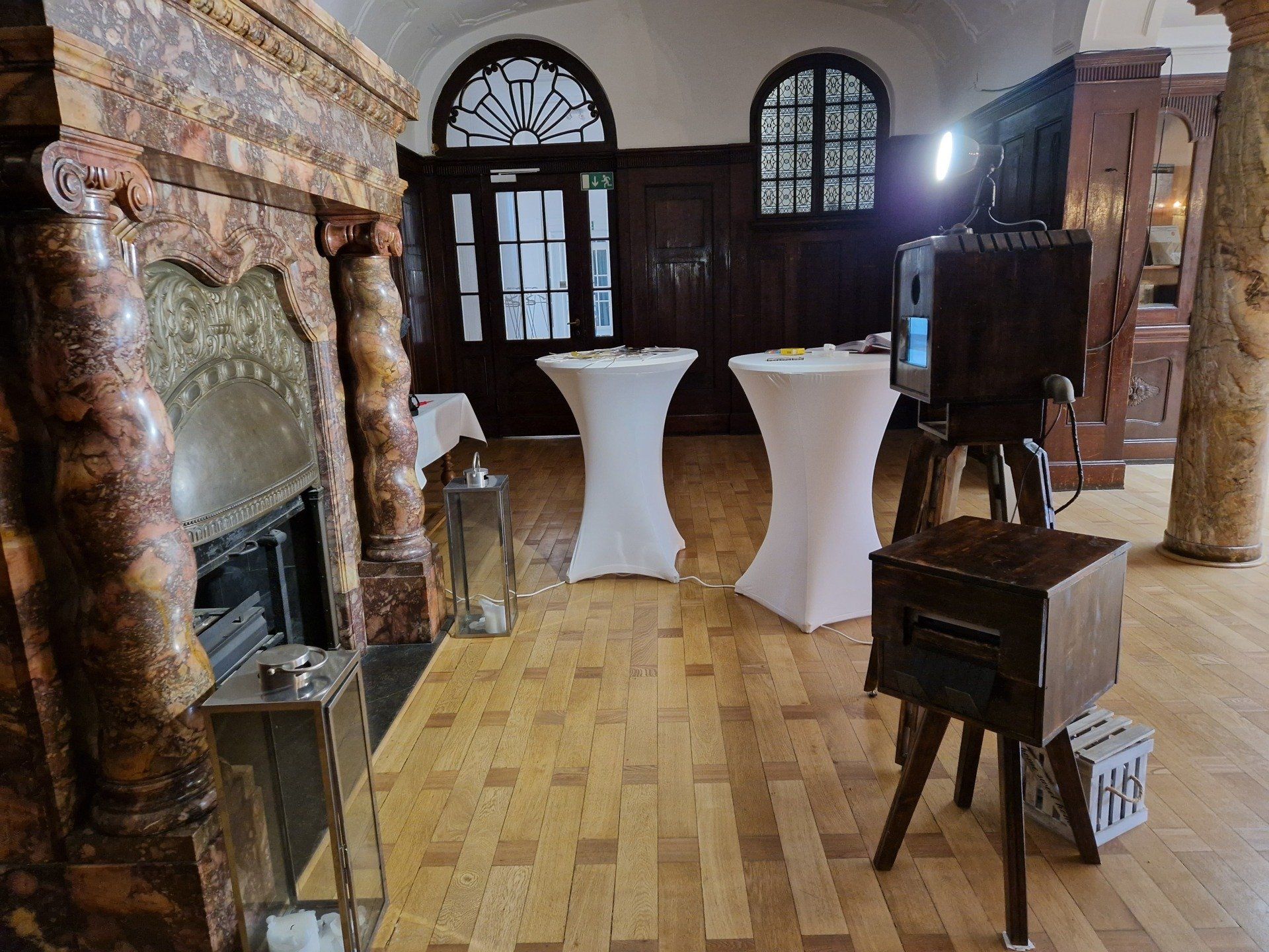 Ein Zimmer mit Kamin, zwei weißen Stehtischen und einer nostalgischen Fotokabine auf Holzboden.