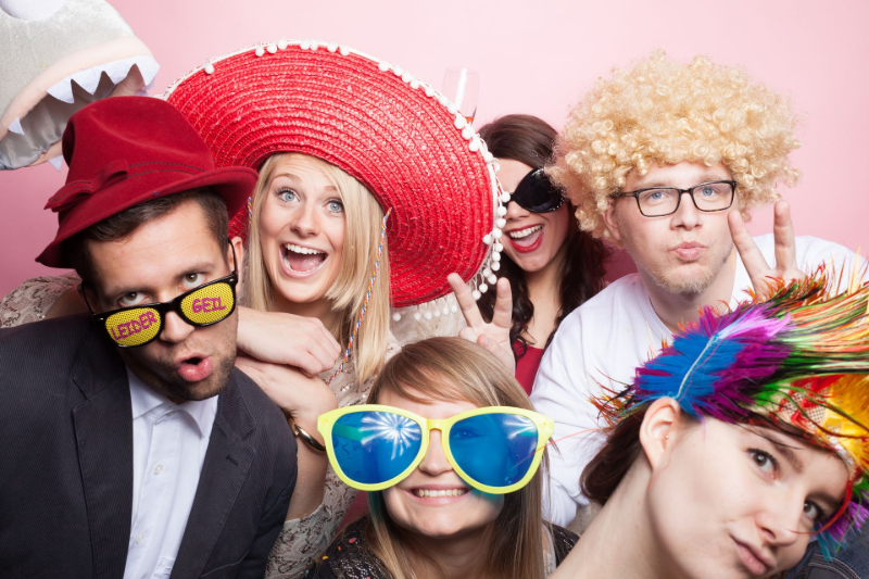 https://stock.adobe.com/de/search?k=fotobox&search_type=usertyped&asset_id=101585279 Eine Gruppe von Menschen in einer Fotokabine, die lustige Hüte und Brillen tragen und mit albernen Gesichtsausdrücken vor einem rosa Hintergrund posieren.