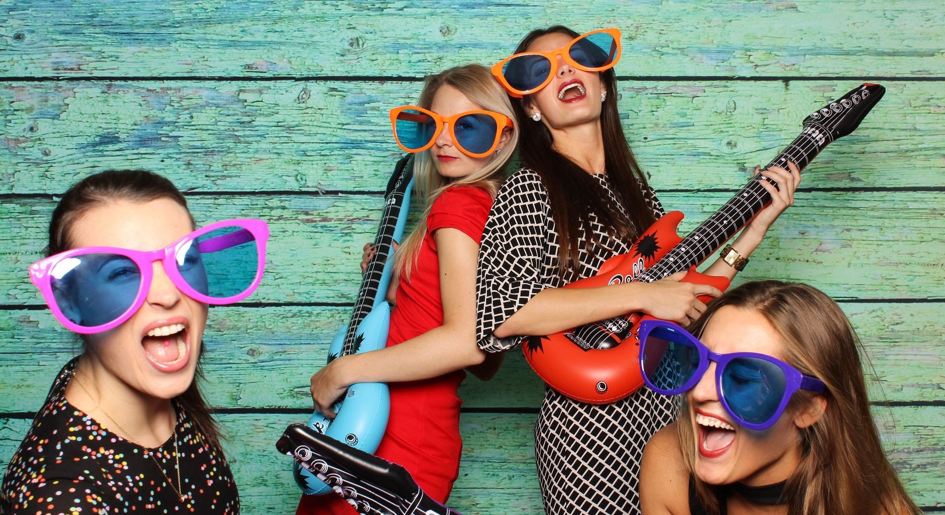 Vier Personen mit übergroßen Sonnenbrillen posieren mit Spielzeuggitarren vor einem türkisfarbenen Holzhintergrund.