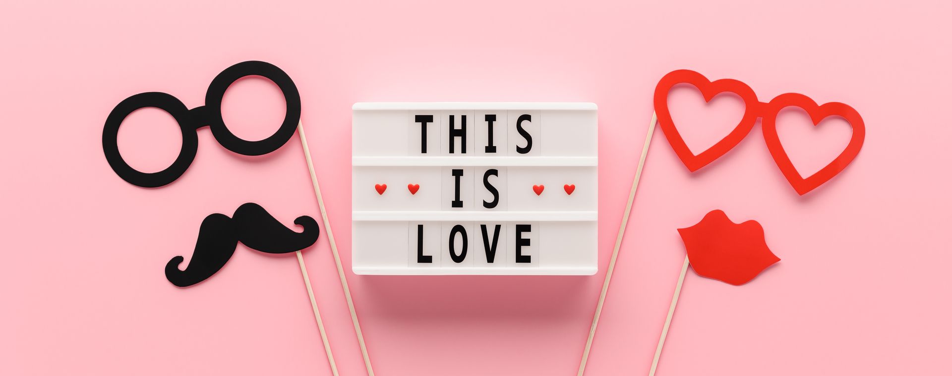 Pink background with props of glasses, mustache, hearts, lips, and a lightbox that reads “THIS IS LOVE”.