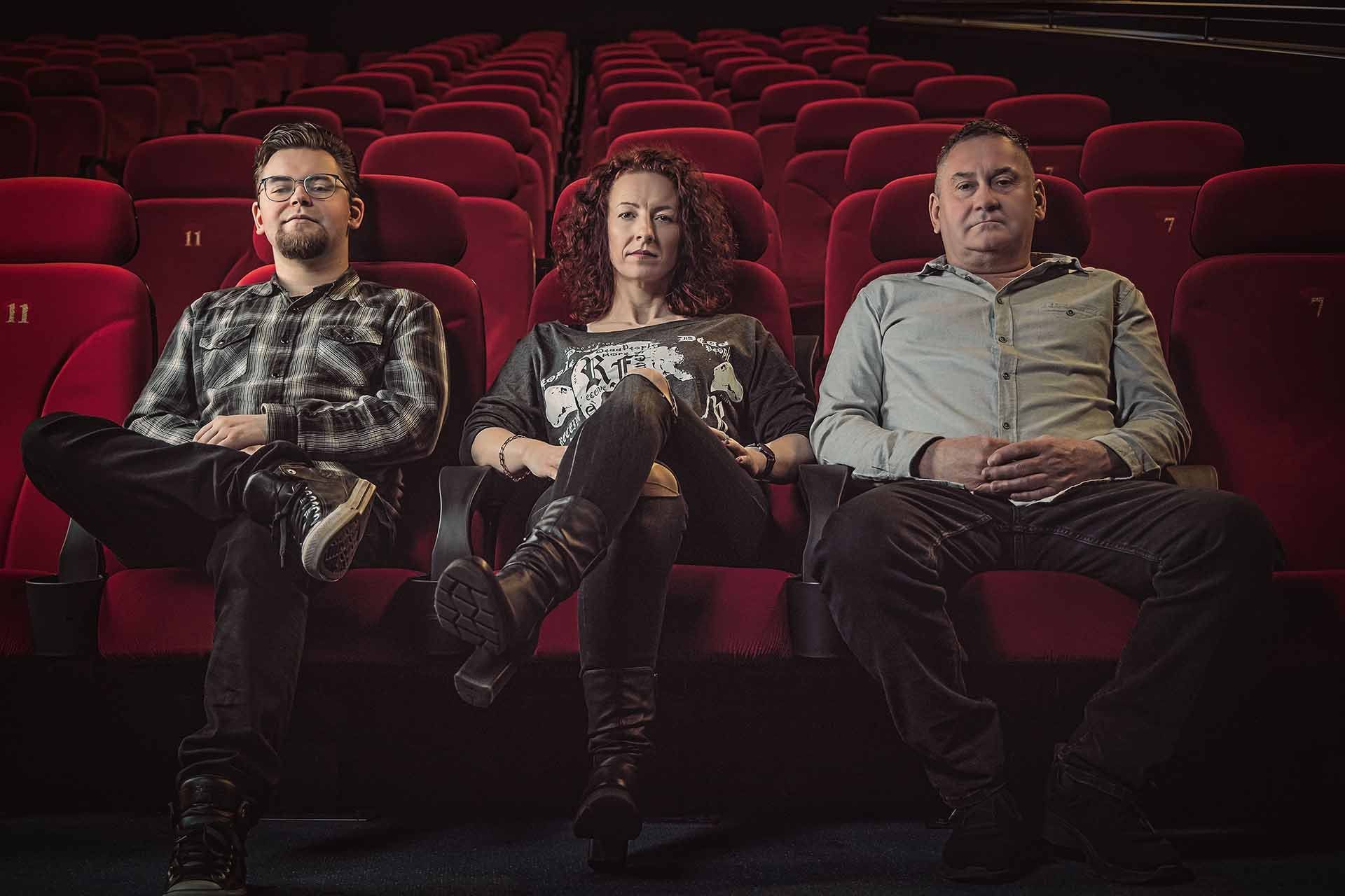 Drei Personen sitzen in roten Sitzen in einem Theater.